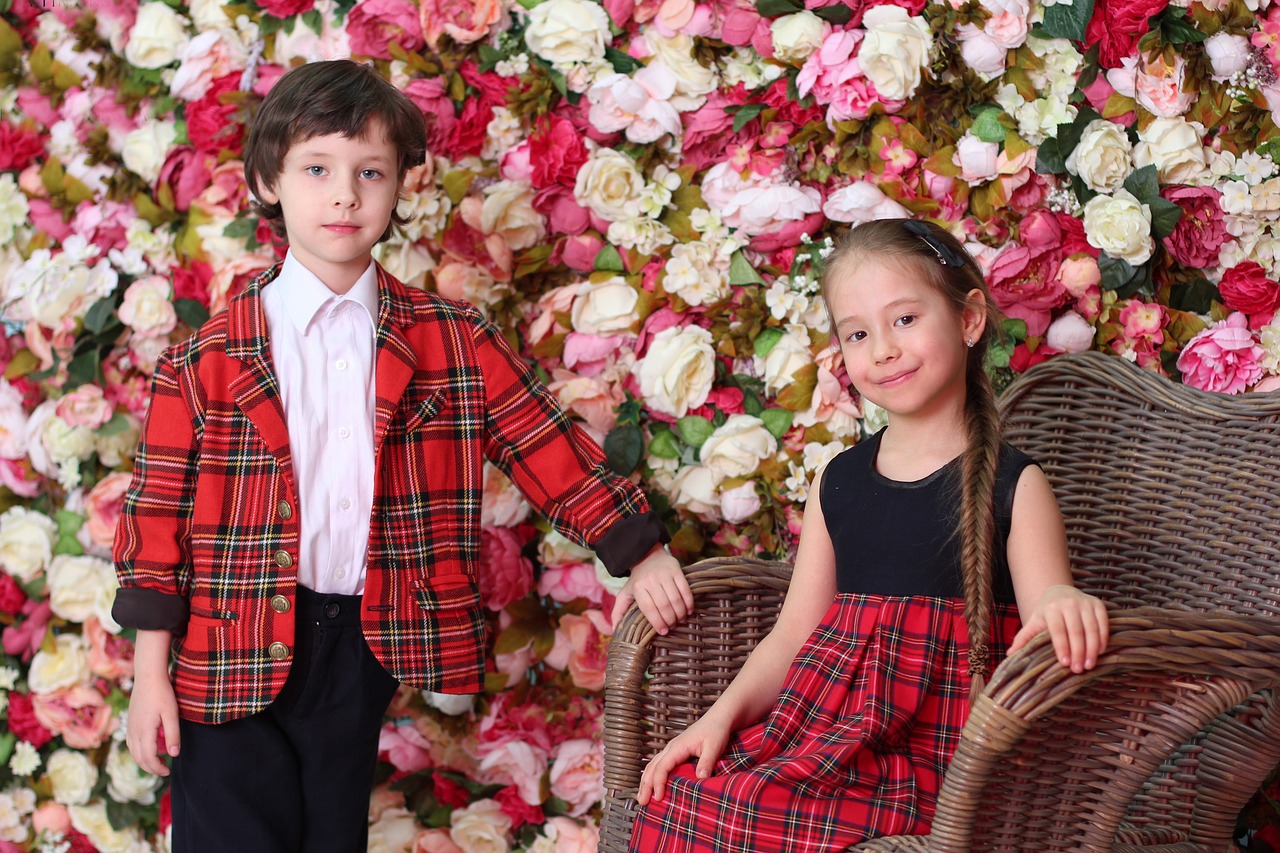 赤のタータンチェックのお洋服を着ている可愛い男の子と女の子のお写真。後ろには白・ピンク・赤のバラのお花が一面に咲いている。